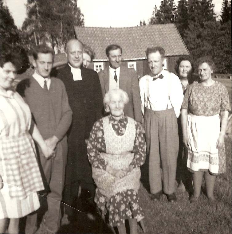 Gruppfoto med åtta personer och en präst framför ett trähus