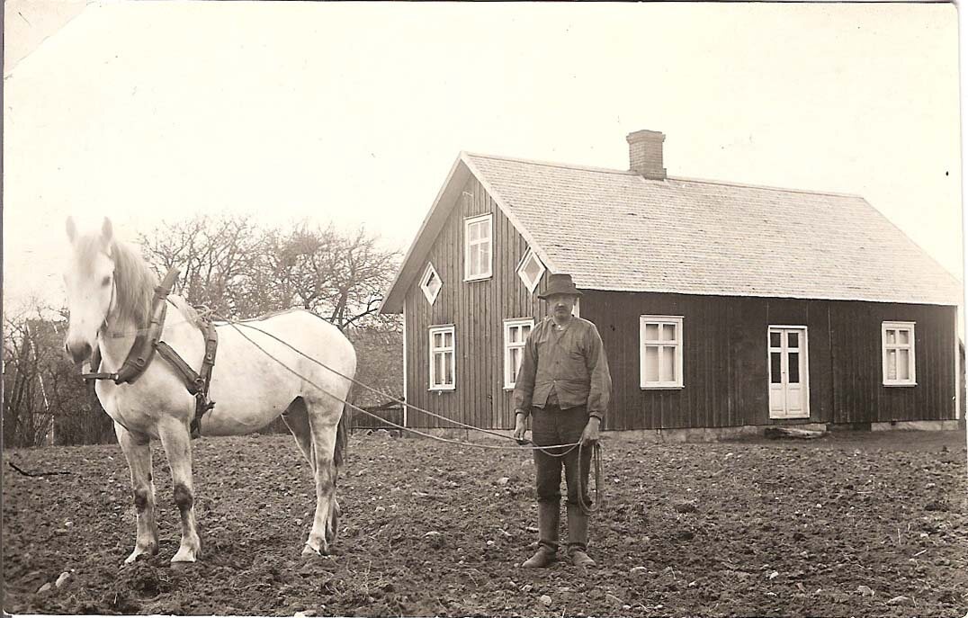 Lantbrukare med arbetshäst på en åker framför ett trähus